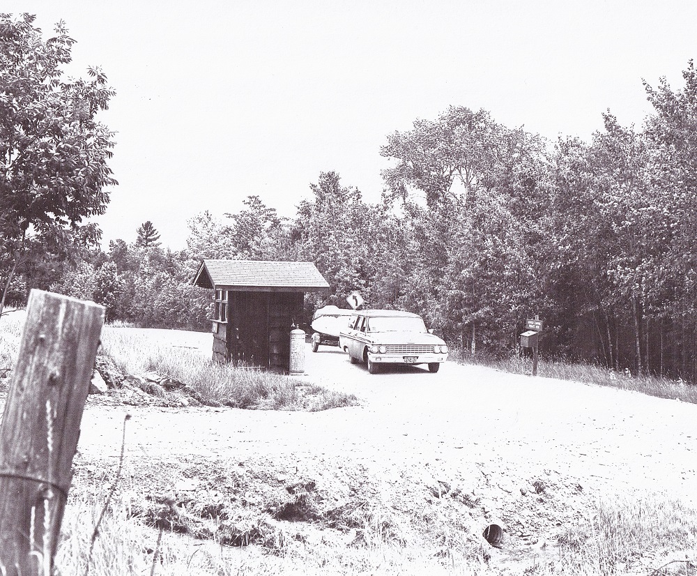 Black-and-white image of vintage vehicle towing boat trailer on dirt road past shack with trees in background