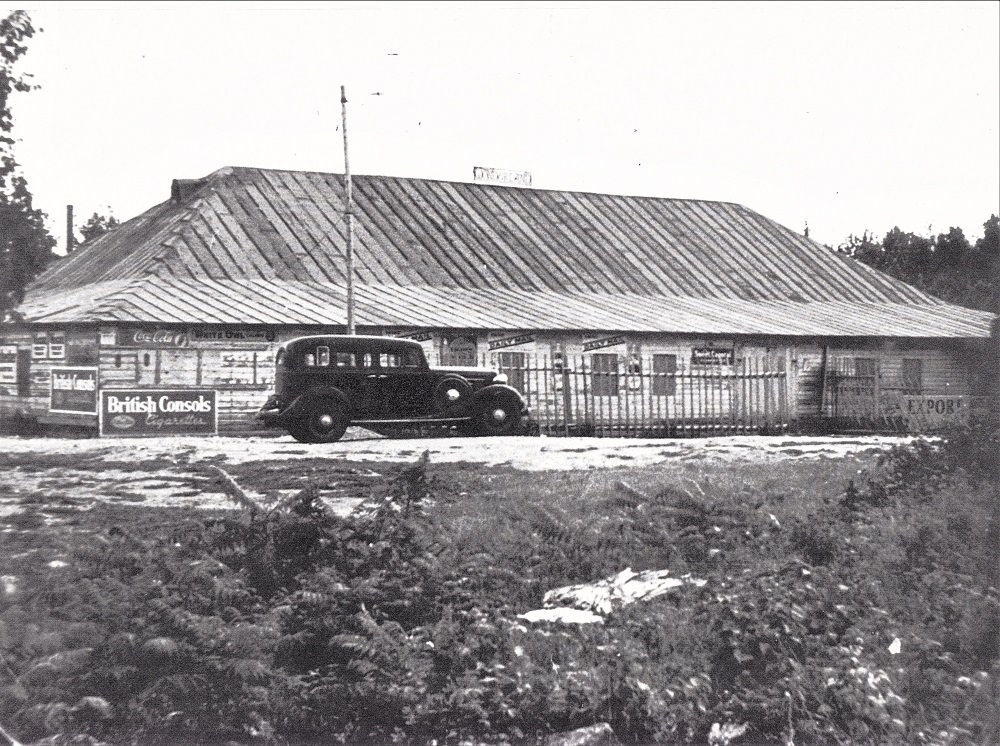 Black-and-white image of large vintage car parked in front of building with enormous wooden roof