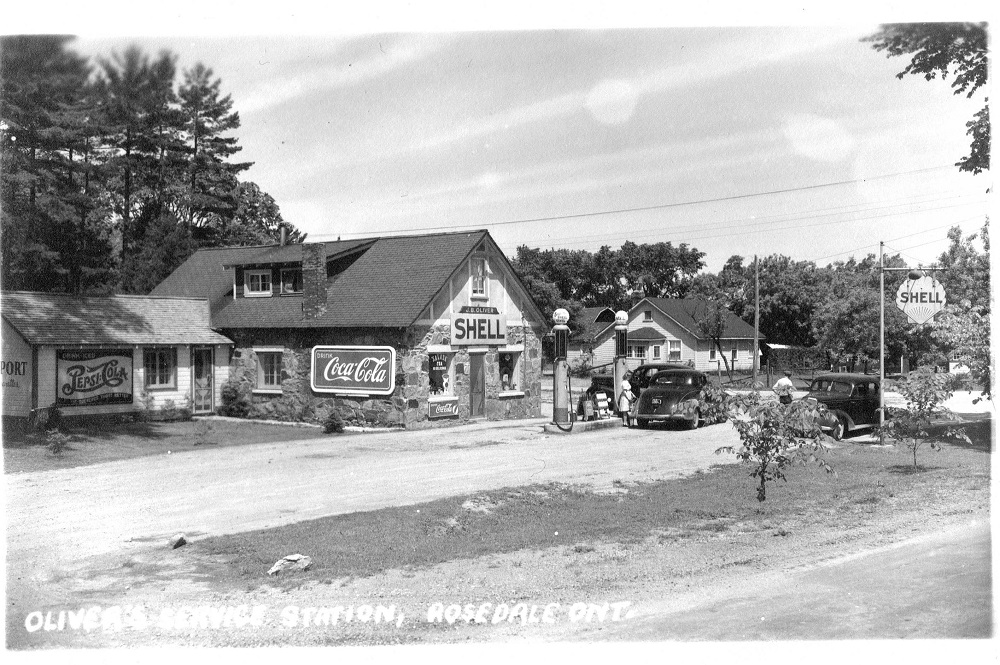 Black-and-white image of vintage cars parked in front of gas pumps with stone building in background