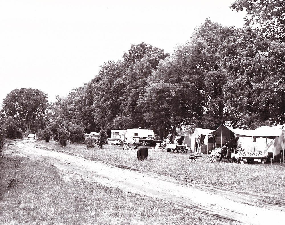 Black-and-white image of tents and trailers parked beneath trees, surrounded by camping supplies, with muddy road in foreground
