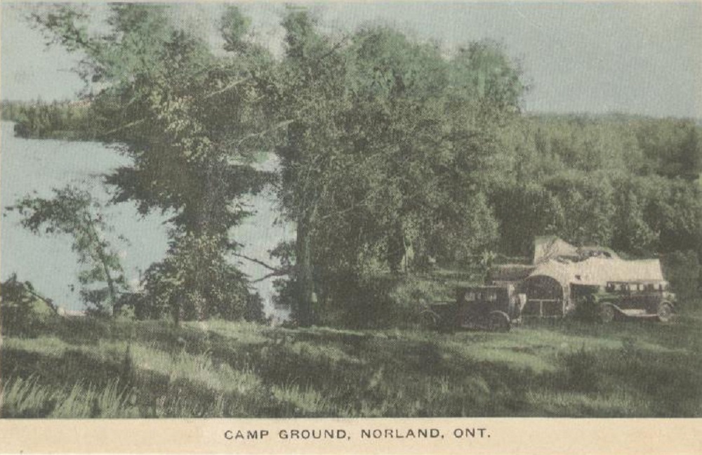 Colour image of vintage vehicles and white tents beneath trees with lake in background