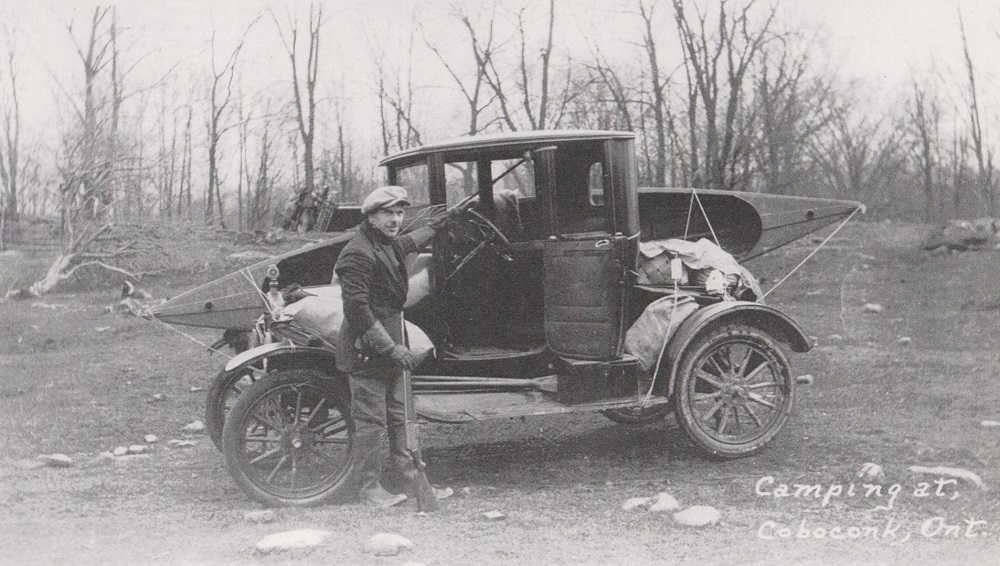 Black-and-white image of man beside vintage vehicle carrying canoe and camping gear with trees in background