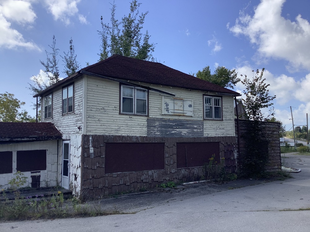 Colour image of abandoned building with weeds and asphalt in foreground and trees and sky in background