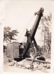 Black-and-white image of steam shovel digging at roadside with trees and hydro poles in background