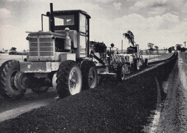 Black-and-white image of machinery and loose asphalt on highway with trees in background