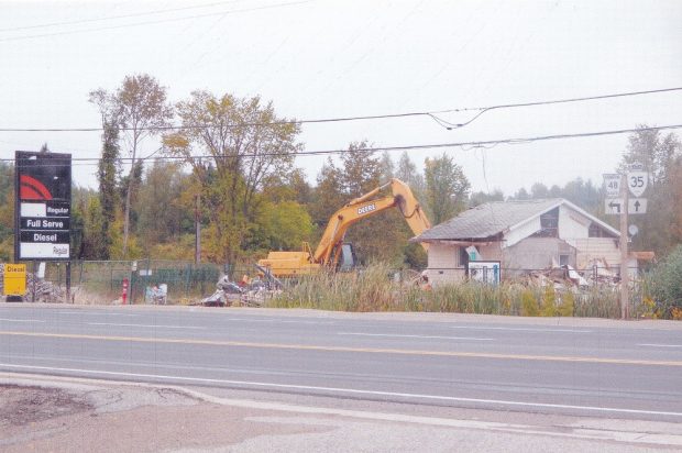 Colour image of machine knocking down a building with trees in background and sign and highway in foreground