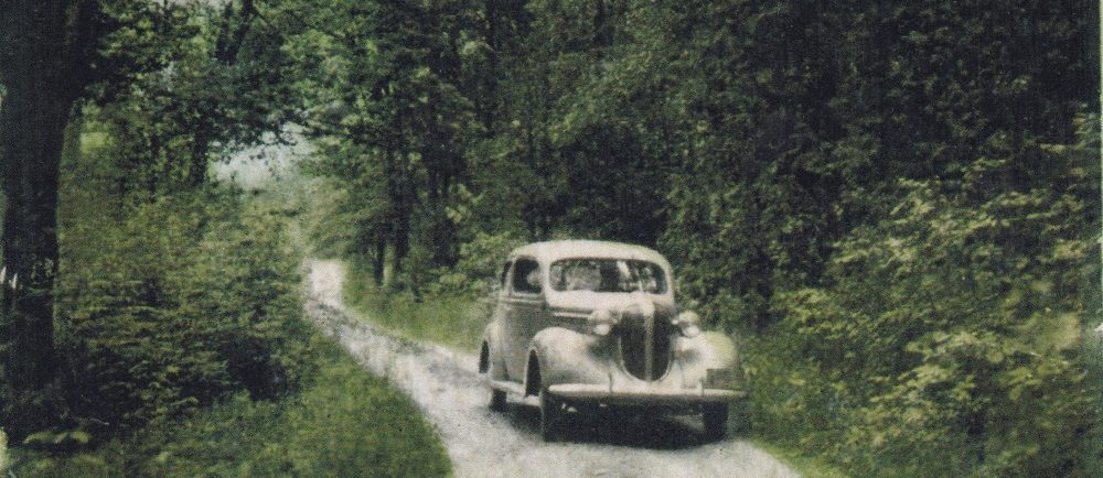 Colour image of a vintage vehicle travelling along road with trees and foliage on either side