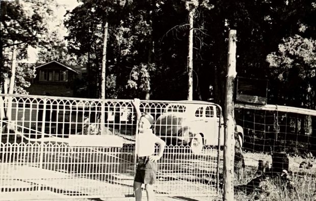 Black-and-white image of child standing beside metal gate with building, trees, and vintage car in background