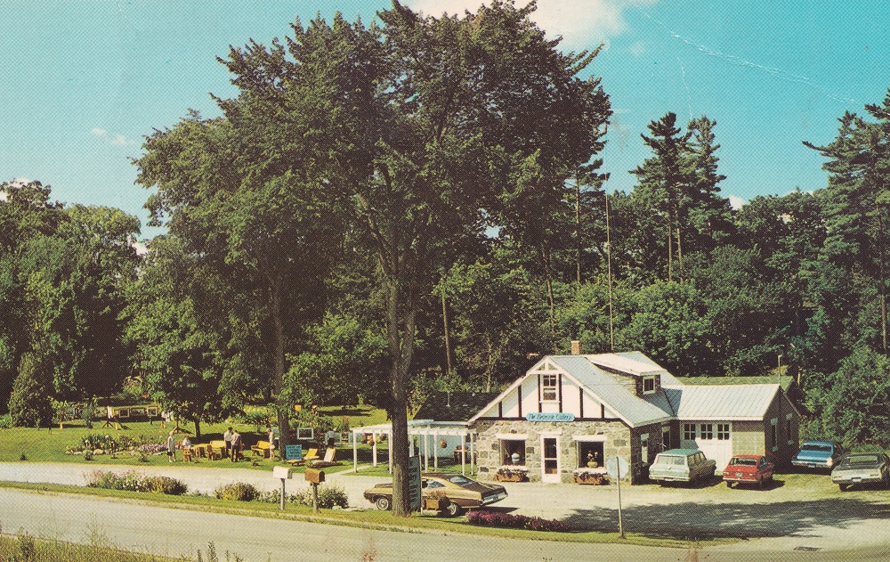 Colour image of stone building surrounded by gardens and trees with cars and parking lot in foreground