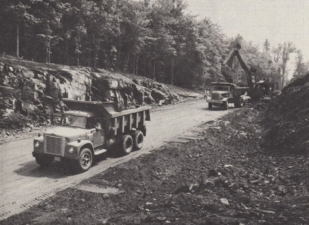 Black-and-white image of dump trucks moving earth on road with trees in background