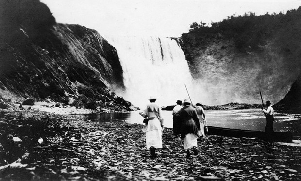 Silver print of Elsie Refird, Evelyn MacInnes walking on the stones approaching the base of the falls of the Metis River and about to embark in the canoes for a day of fishing