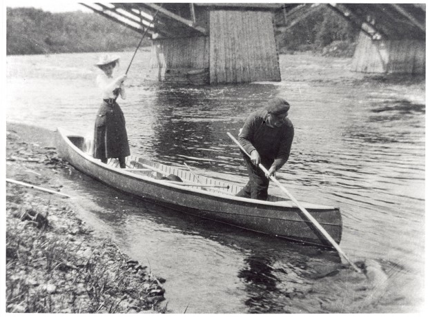 Silver print showing Elsie Reford standing in a canoe at the river's edge. She is wearing a mid-length skirt, a white blouse, and a large elegant hat. She appears to be pulling on a large fishing pole. A man at the front of the canoe is lifting a large fish from the river with a long pole and hook.