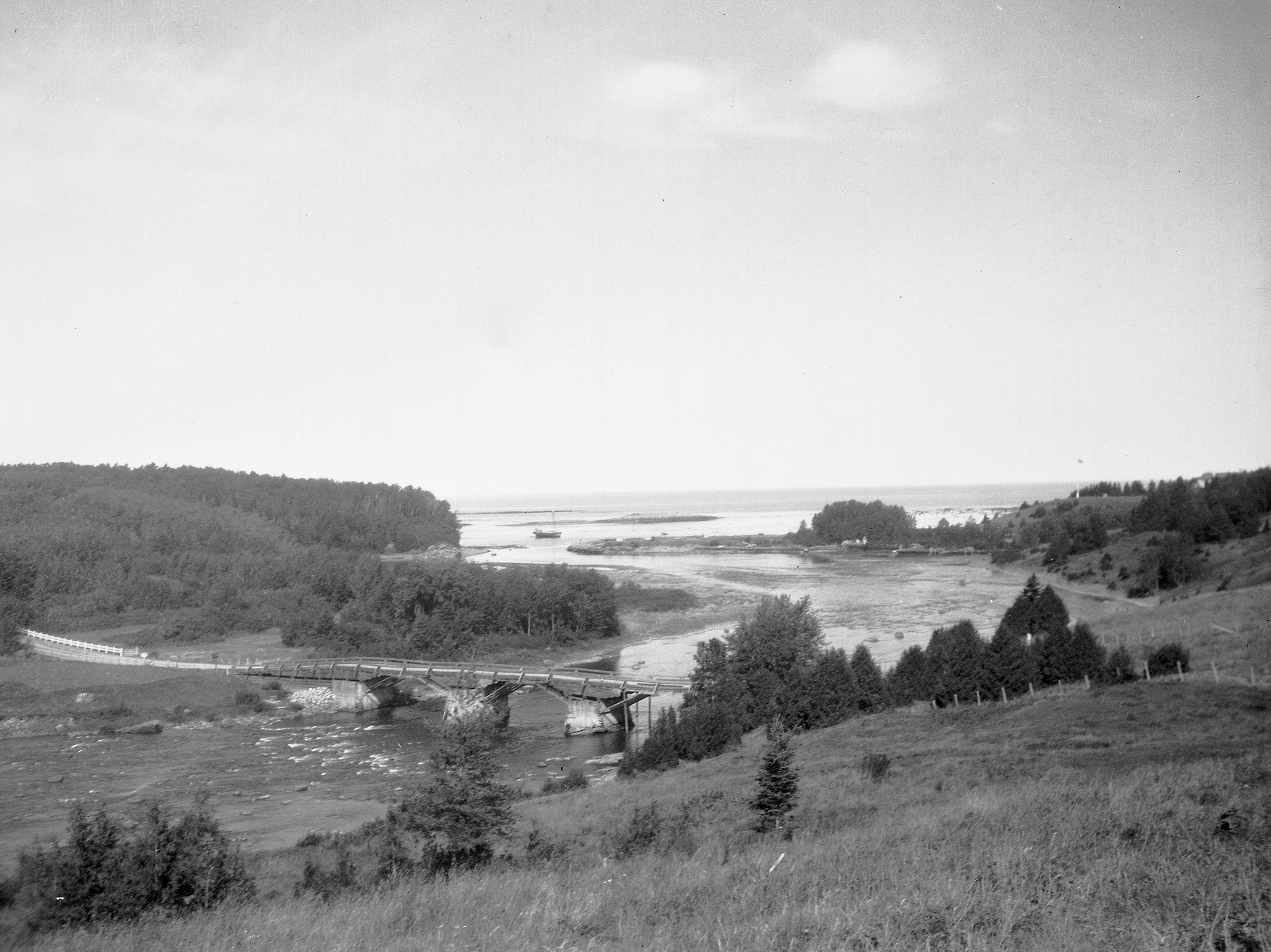 View of the mouth of the Metis River showing the old wooden bridge, the remains of the wharf of the Price Brothers Company, a schooner and the mast at the belvedere of Estevan Lodge.