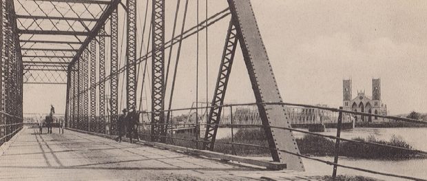View of Sainte-Anne-de-la-Pérade’s bridge spanning the Sainte Anne River and on which there is a horse-drawn carriage led by two men and there are also two other men leaning on the railing. In the distance we catch sight of Sainte-Anne Church.