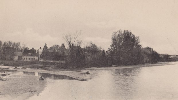 Black and white photograph of John Jones Ross’s house on île Saint-Ignace taken from the municipal bridge and of which the roof sticks out of the trees.