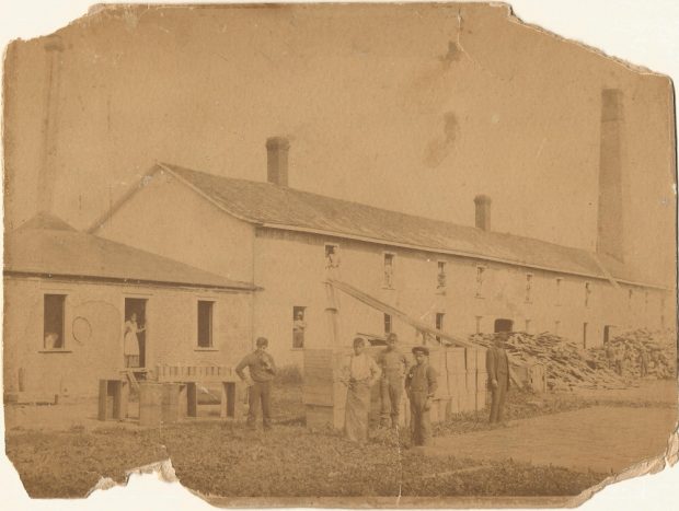 Archival photograph of the building housing the match factory in front of which several workers stand. Piles of wooden debris border the frontage which has many windows where men and women workers can be seen.
