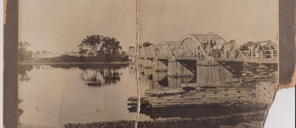 Black and white photograph of Sainte-Anne’s bridge of which two arches of the wooden structure were destroyed by a scree and with debris lying in the river.