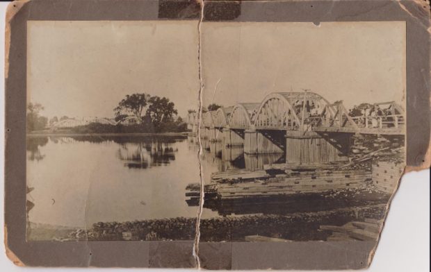 Black and white photograph of Sainte-Anne’s bridge of which two arches of the wooden structure were destroyed by a scree and with debris lying in the river.