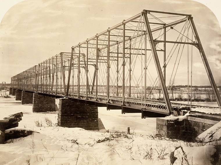 Black and white winter photograph of the railway bridge passing over Sainte-Anne River with its iron structure and stone pillars.