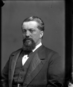 Photographie en noir et blanc de John Jones Ross portant la barbe et habillé d’un complet avec nœud papillon.