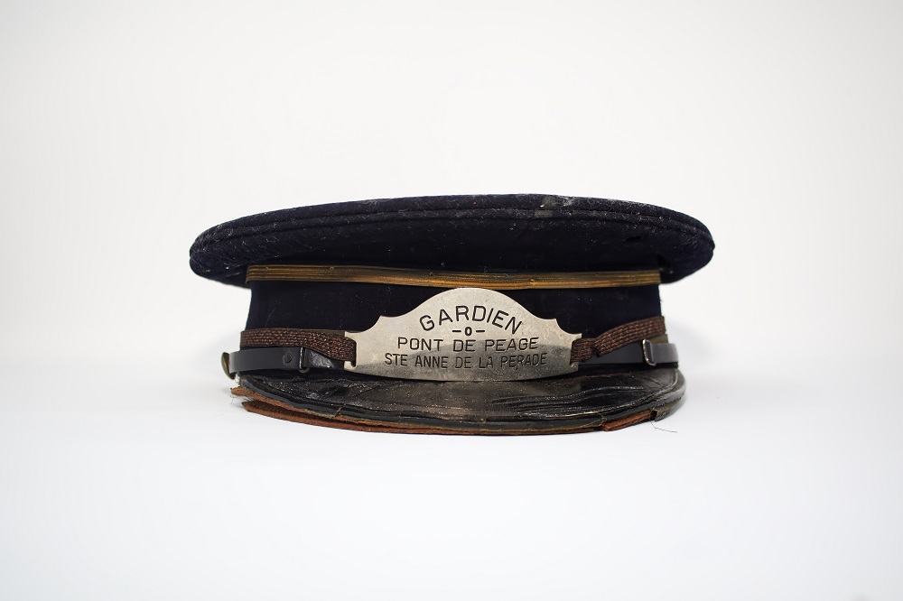 Front view of a black cap with golden lace with the inscription Sainte Anne de la Pérade Toll bridge keeper carved on a metal badge.