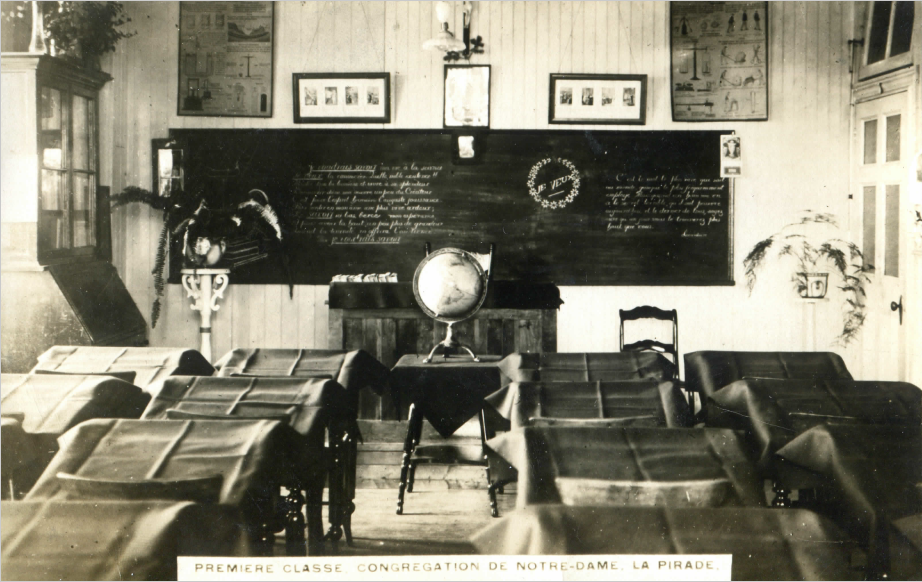 Photographie noir et blanc d’une classe du couvent de Sainte-Anne avec tableau noir globe terrestre et pupitres recouverts de draps.