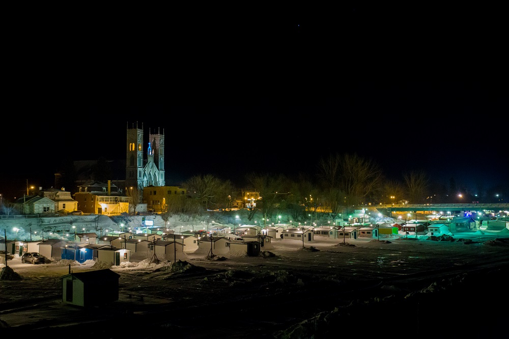Night photograph of dozens of fishermen’s cabins on the Sainte-Anne River in front of the illuminated church.