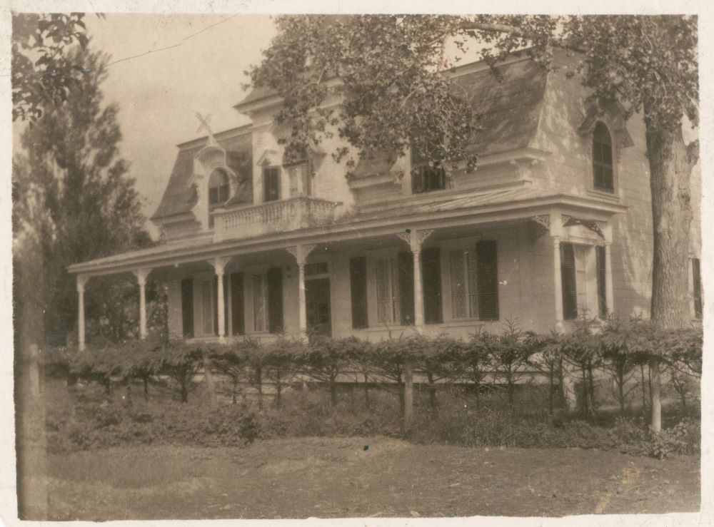 Black and white photograph of the front view and part of the side of J A Rousseau’s house Second Empire style with arched dormers and covered porch.