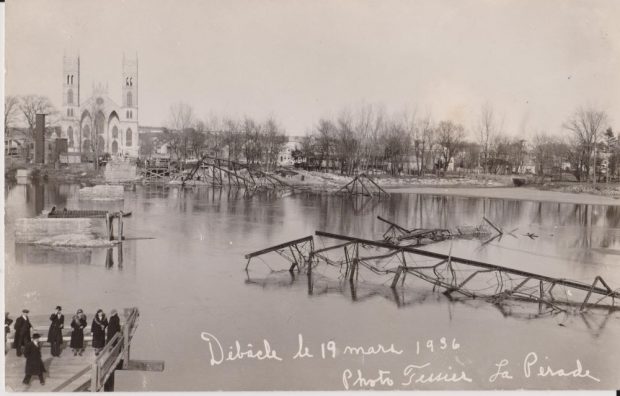 Black and white photograph of the iron bridge destroyed by the ice breakup of March 19 1936 with the structures remaining in the river and the Sainte-Anne Church on one shore. A gathering of seven people observes the damage from the remains of the footbridge on the other shore.