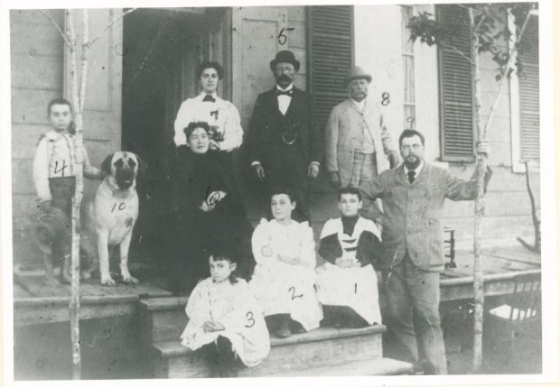 Black and white photograph of Jeffrey Alexandre Rousseau his wife his four children and three other family members with a dog posing in front of his house’s porch.