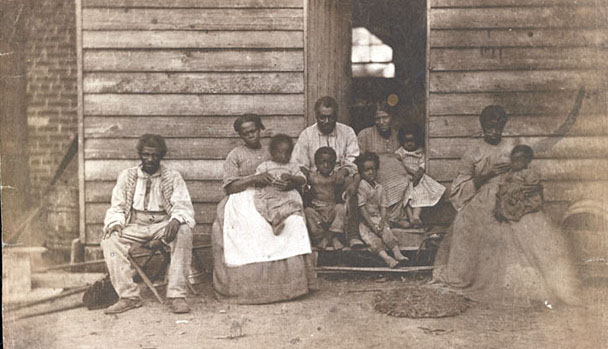 A family of slaves is photographed in front of the ramshackle hut where they live