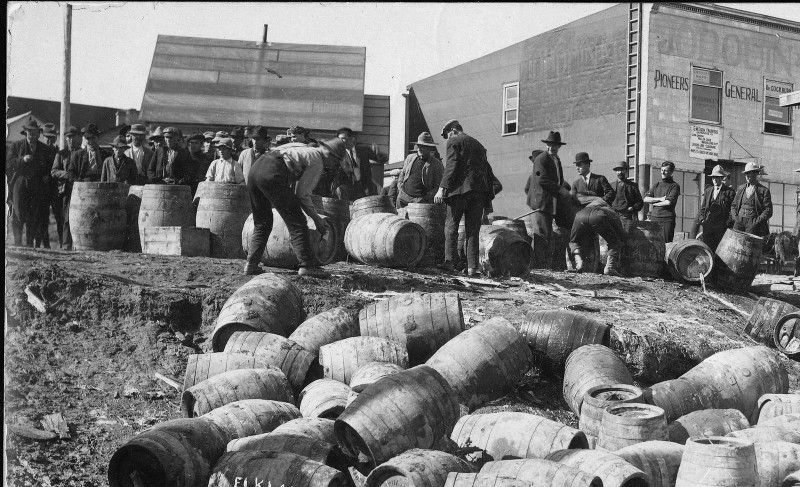 Workers rip open barrels to dump the alcohol; the empty barrels pile up along a gentle slope.