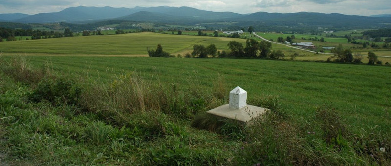 In the middle of a leafy field at the foot of the mountains, a small white marker indicates the Canada-U.S. border.