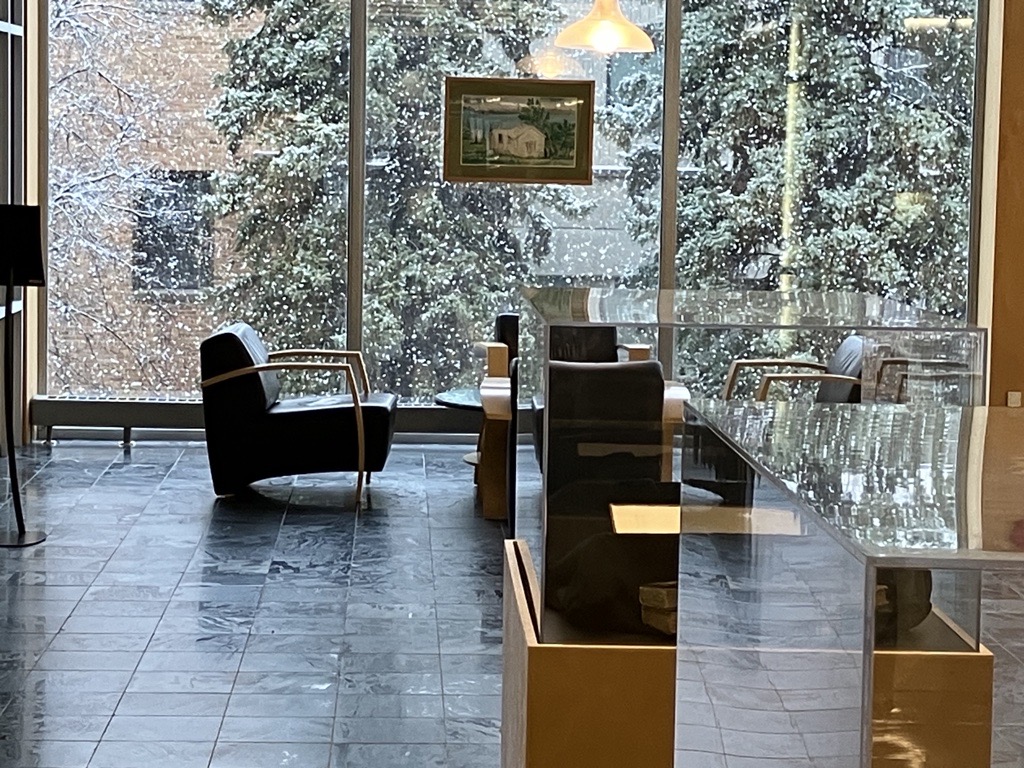 A quiet reading room with large floor-to-ceiling windows revealing a snowy outdoor scene with tall evergreen trees. Inside, modern black chairs with curved armrests face the window. Display cases with books or artifacts line the room, and soft natural light illuminates the space. A framed artwork hangs in the center of the window.