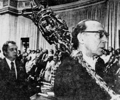 Black and white photograph of a ceremonial procession in a formal chamber. A man in glasses leads the procession, holding an ornate, dragon-headed ceremonial staff. Behind him, another man in a suit follows. Rows of people in formal attire are seated and standing in the background, with large curtains and classical architectural features framing the scene.