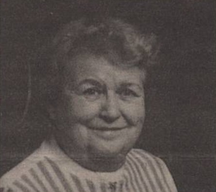 Black-and-white portrait of an elderly woman with short, curled hair. She is smiling softly and wearing a light-colored blouse with vertical stripes.