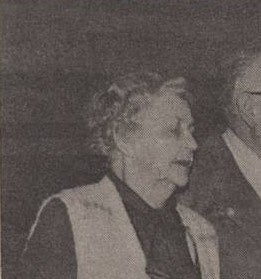 Photo granuleuse en noir et blanc d’une femme âgée avec les cheveux courts pis frisés, tournée vers la gauche. Elle porte un gilet pâle par-dessus une blouse foncée pis on dirait qu’elle est en train de parler. On voit une partie d’un homme en complet à sa droite. L’arrière-plan est sombre pis pas distinct.