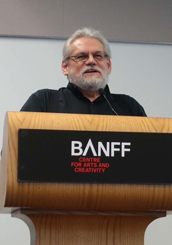 An older man with white hair, beard, and glasses speaks at a podium with a microphone. The podium has a sign that reads BANFF Centre for Arts and Creativity.