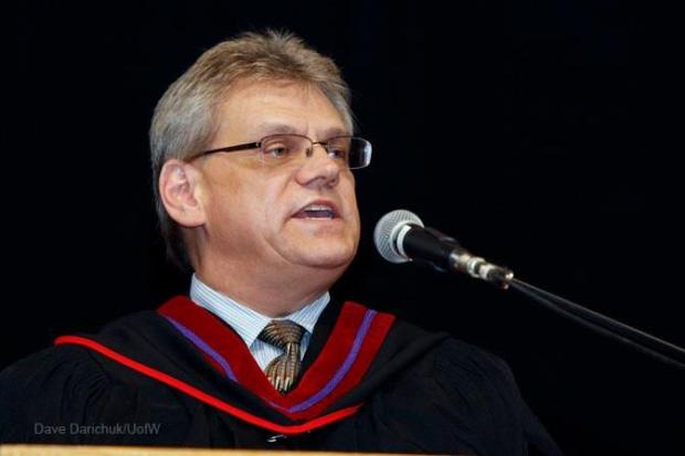 Un monsieur avec les cheveux gris courts pis des lunettes parle dans un micro pendant une cérémonie officielle. Il est habillé d’une chemise pis d’une cravate.