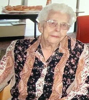 Aldrað kona með hvítt hár og gleraugu situr inni. Hún er klædd mynstraðri blússu með blóma- og kasmíramynstri í dökkum og ljósum tónum. Fyrir aftan hana er borð með bakka af bakkelsi og gluggi sem hleypir inn náttúrulegu ljósi.