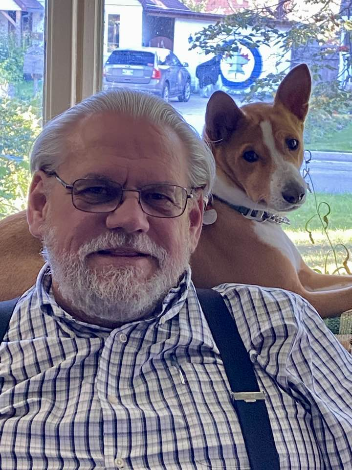 An older man with glasses, white hair, and a beard sits indoors in front of a window, wearing a plaid shirt with suspenders. A brown and white dog rests on the back of the couch behind him, looking toward the camera.