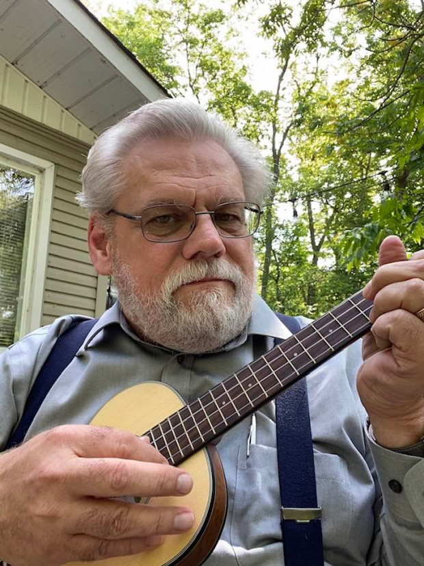 Un monsieur plus âgé avec les cheveux blancs, une barbe pis des lunettes est assis dehors en train de jouer d’un petit instrument à cordes. Il porte une chemise grise avec des bretelles, pis y’a des arbres partout en arrière.