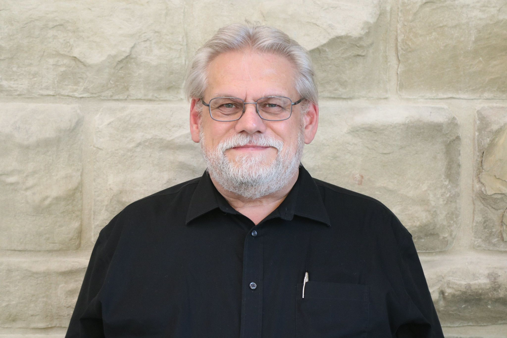 An older man with white hair, beard, and glasses stands in front of a stone wall, wearing a black collared shirt with a pen in the breast pocket.
