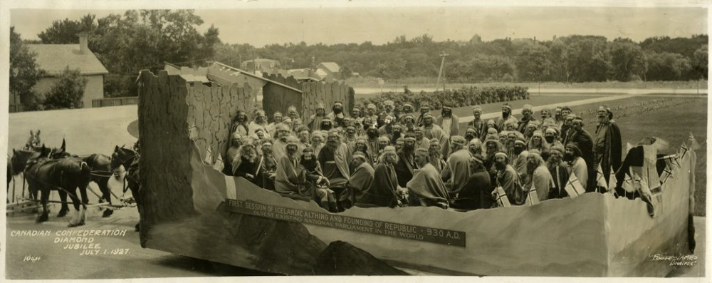 Photo en noir et blanc d’un grand char allégorique datant du 1er juillet 1927, avec des dizaines de personnes déguisées en premiers colons islandais. Le char est en forme de grande salle de rassemblement viking et est tiré par des chevaux. Une bannière sur le côté indique : Première séance de l’Althing islandais et fondation de la République, 930 apr. J.-C. – Le plus ancien parlement national existant au monde. La légende de l’image : Jubilé de diamant de la Confédération canadienne, 1er juillet 1927. 