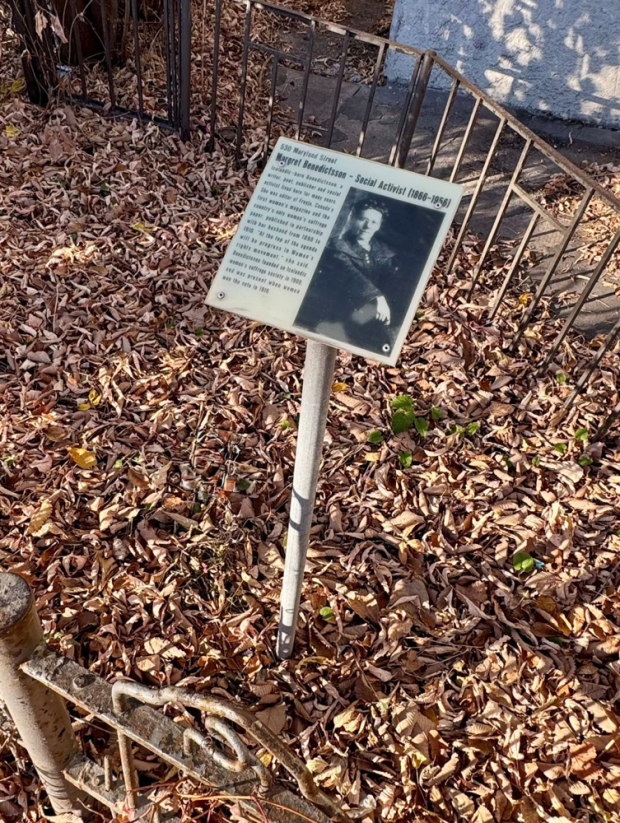 Outdoor informational sign mounted on a metal post, standing among dry fallen leaves within a fenced area. The sign includes a photograph of a man dressed in dark clothing along with a block of text providing historical context. Behind the sign are metal railings and a light-colored structure partially visible in the background. The area is shaded with filtered autumn sunlight.