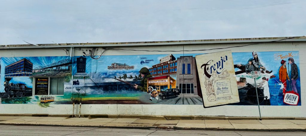 A wide mural painted on a brick wall showing historical scenes from Winnipeg’s West End. The artwork includes images of trains, a bridge, factories, wartime soldiers, and iconic local businesses like Safeway and Christie's Biscuits. Prominently featured is a large painted cover of Freyja, a women’s and suffrage newspaper, along with modern fashion scenes and industrial logos. The mural blends vibrant colors and historical details, highlighting the area’s diverse heritage.