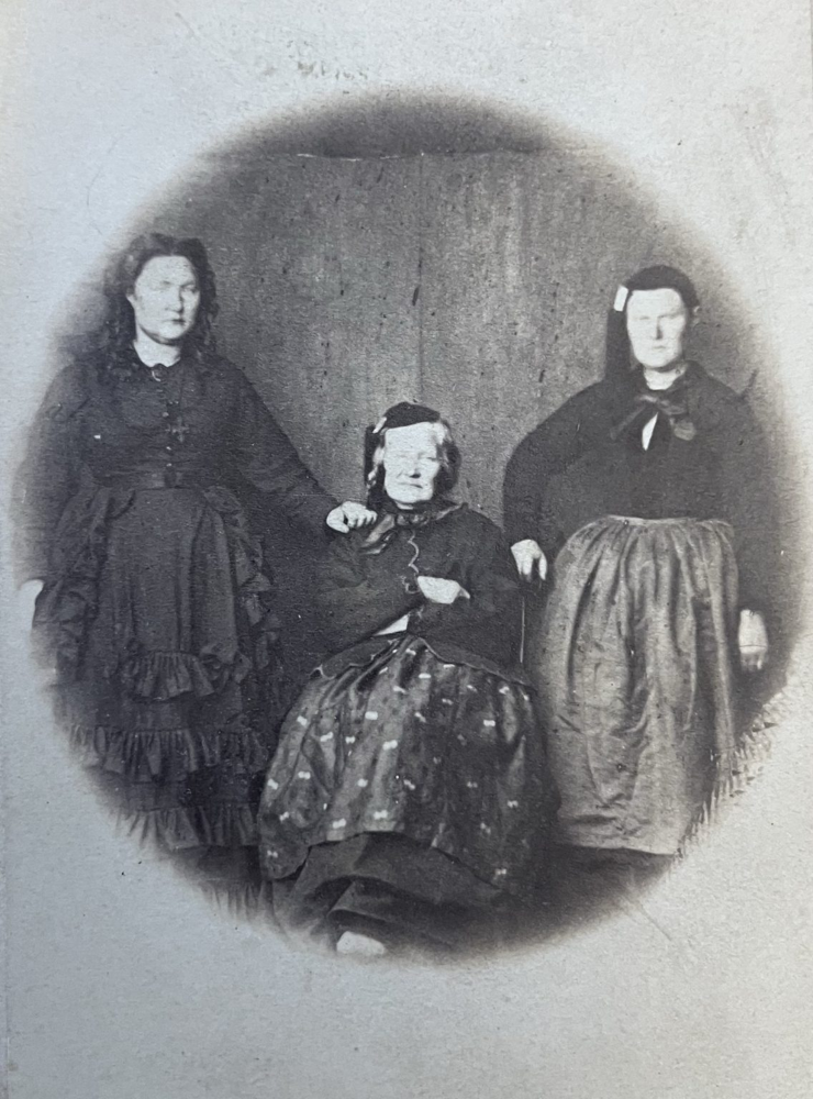 A sepia-toned historical portrait showing three Icelandic women. Torfhildur Þorsteinsdóttir Holm stands on the left in a dark, ruffled dress; her mother Guðríður Torfadóttir sits in the center, wearing a patterned skirt and dark jacket; and her sister Ragnhildur Þorsteinsdóttir stands on the right in a simple blouse and apron. The photo has an oval vignette effect and reflects 19th-century Icelandic attire.