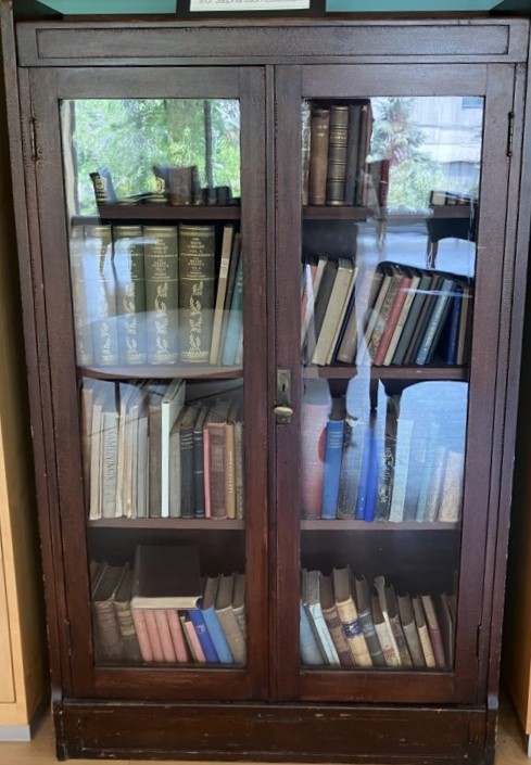 A dark wooden bookcase with glass doors, filled with vintage books in Icelandic and English. The top shelf displays matching gilt-stamped volumes. A small sign on top reads Stephan G. Stephansson Collection. The case stands in a sunlit room with trees visible through a nearby window.