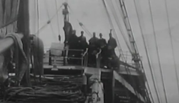 Image en noir et blanc montrant un groupe de personnes debout sur le pont d’un grand voilier, vu d’en arrière de la proue.