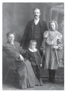 A black-and-white studio portrait of a family. Seated on the left is a woman. Standing behind her is her husband. Their daughter stands to the right in a dress with a lace collar, and their young son stands in front between them. The background features a painted studio backdrop, and all are dressed in formal period clothing.